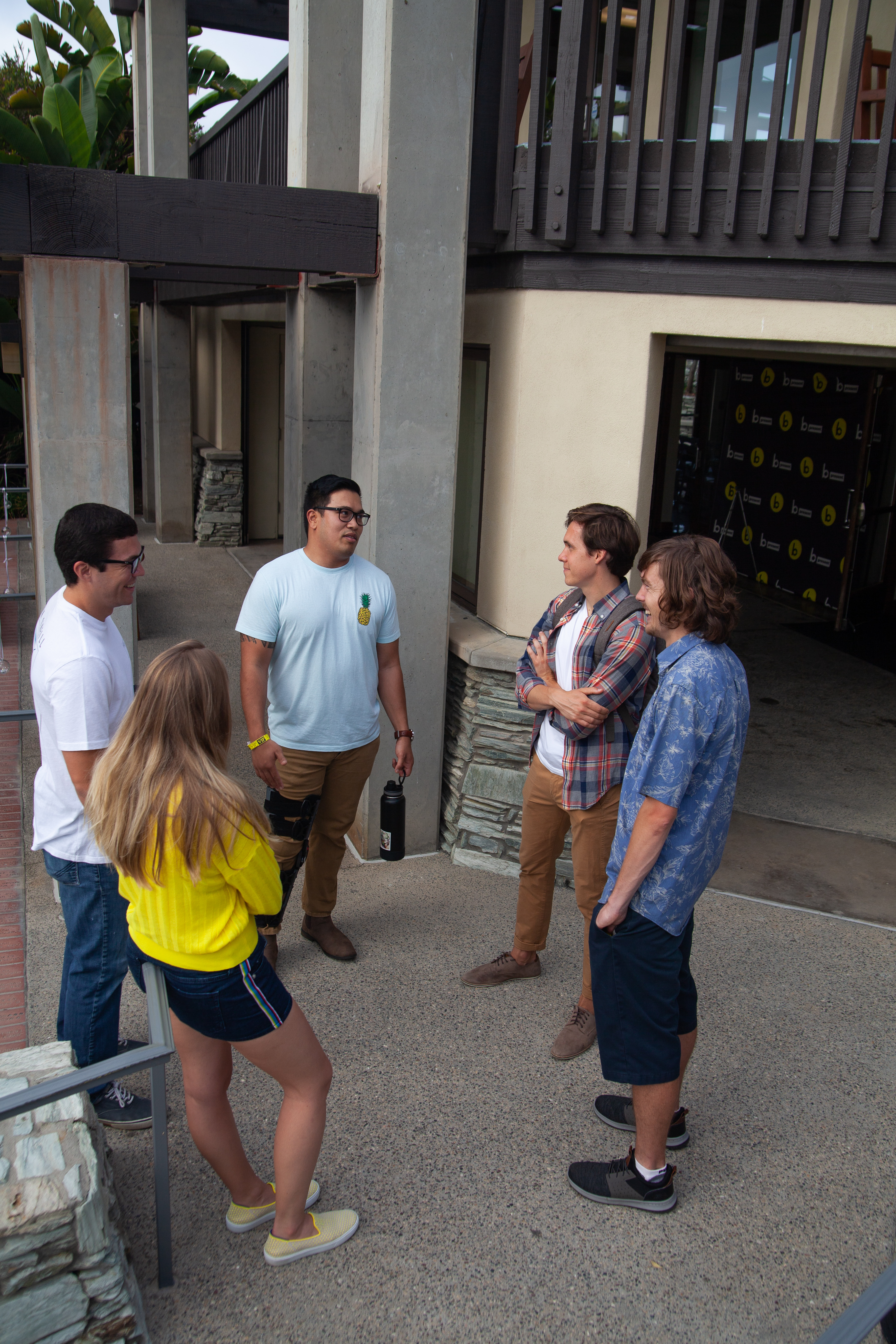 Five young adults stand in a circle outside a building, engaged in conversation. Two women and three men are casually dressed, and the atmosphere appears relaxed and friendly.