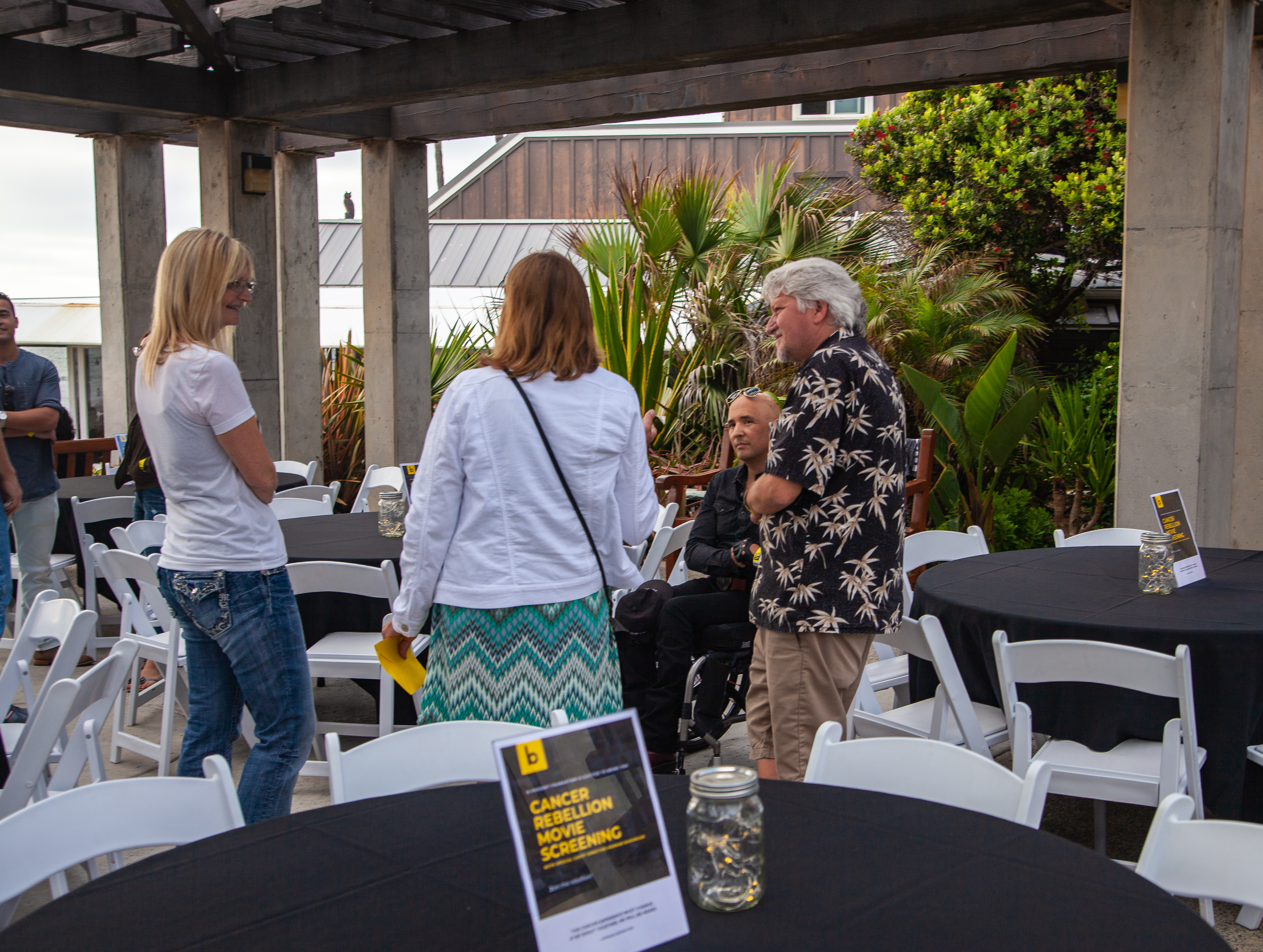 Four people converse by round tables covered with black tablecloths at an outdoor event under a pergola. Signage and jars are on the tables, and palm plants are in the background.