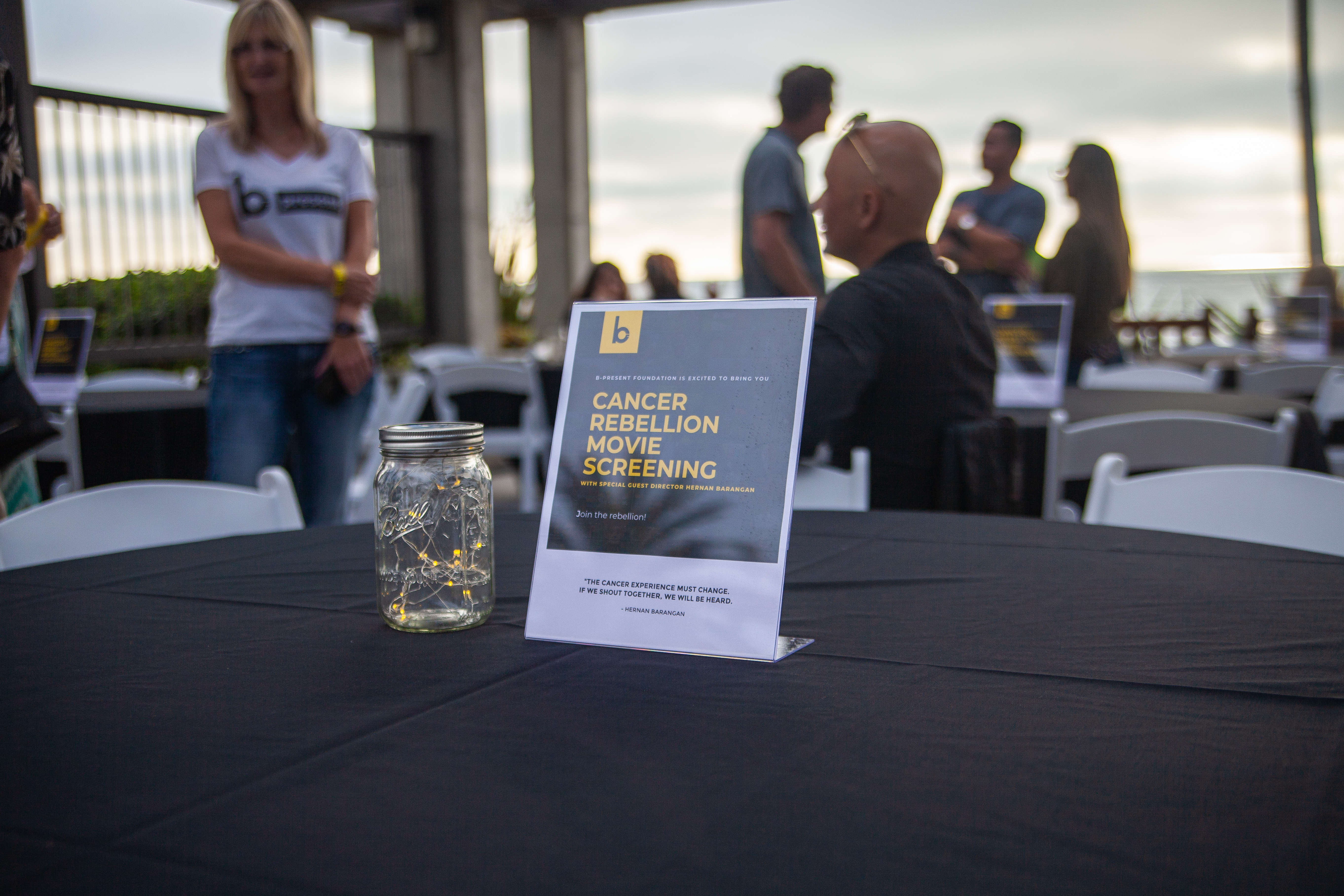 A table at an outdoor event features a sign reading “Cancer Rebellion Movie Screening” and a mason jar filled with string lights. People are seated and standing around other tables in the background.