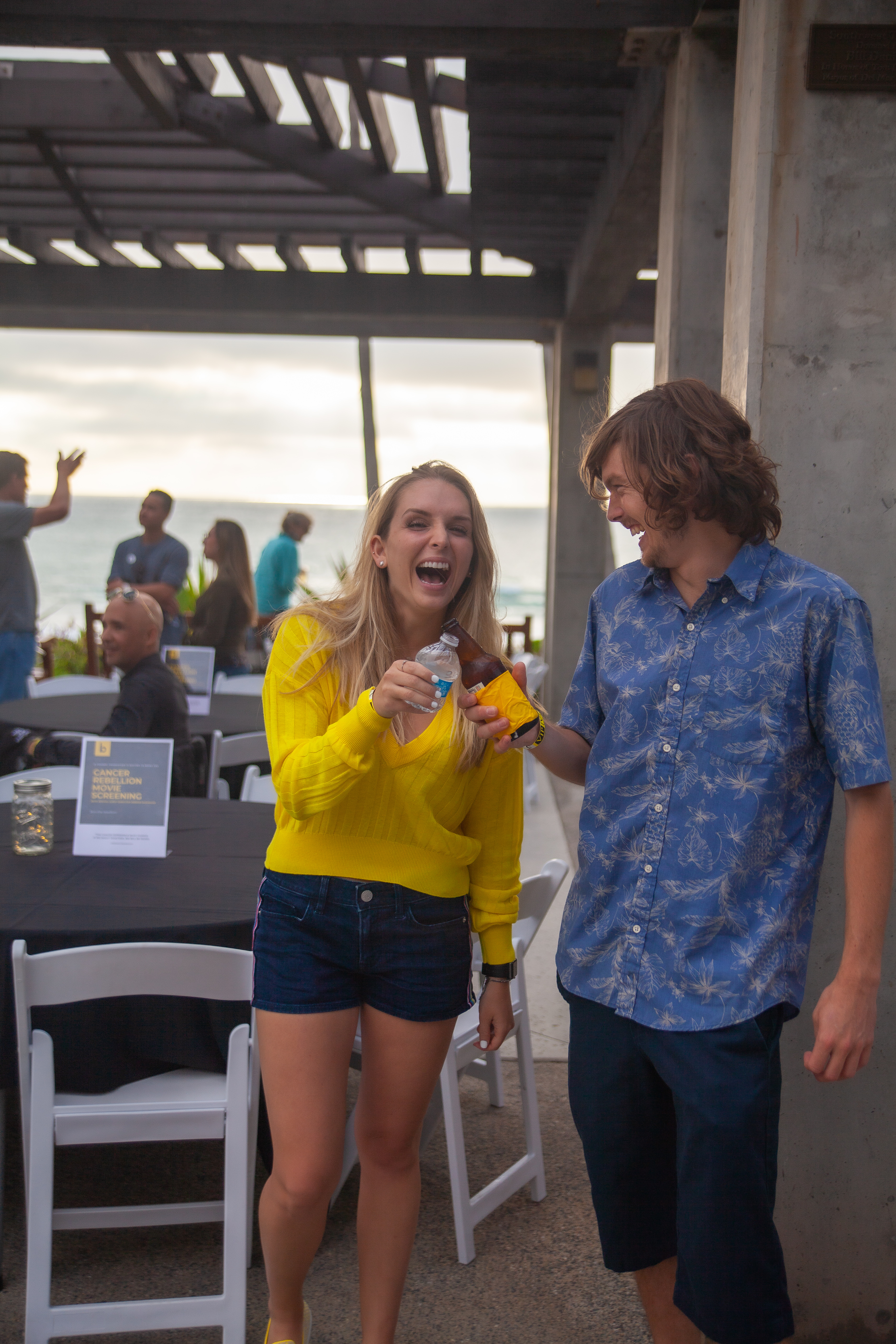 A woman in a yellow sweater and a man in a blue shirt laugh while holding drinks at an outdoor event by the ocean, with other people seated and standing in the background.