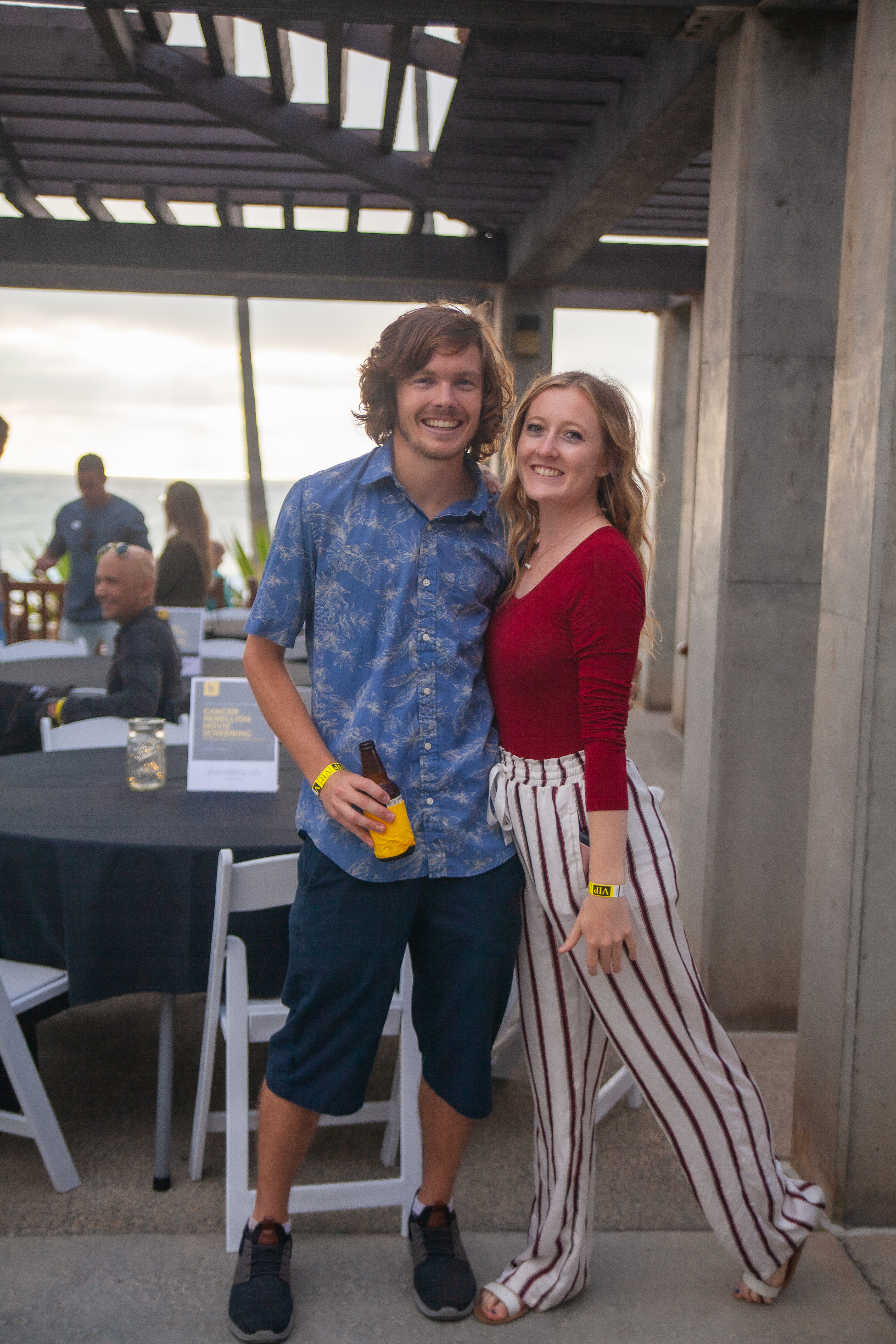 A smiling man in a blue shirt and shorts stands next to a smiling woman in a red top and striped pants. Theyre at an outdoor event with tables and chairs, and the ocean is in the background.