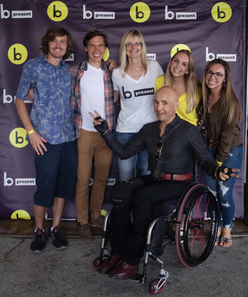 Six smiling people, including one man in a wheelchair, pose together in front of a b-present step-and-repeat banner. The group appears happy and relaxed, with arms around each other.