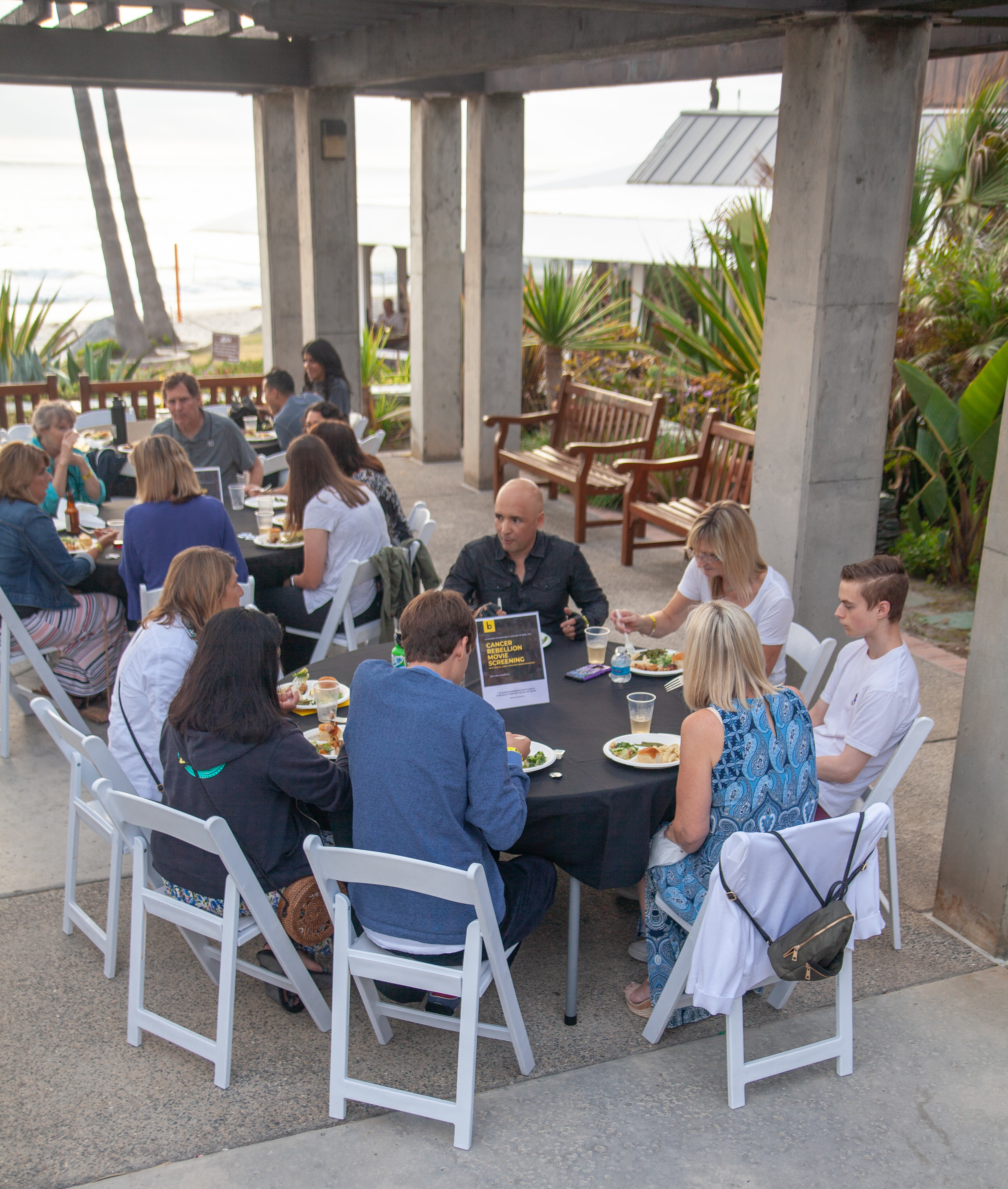 A group of people sit at round tables with white chairs, eating and talking under a pergola near the beach, with greenery and ocean visible in the background.