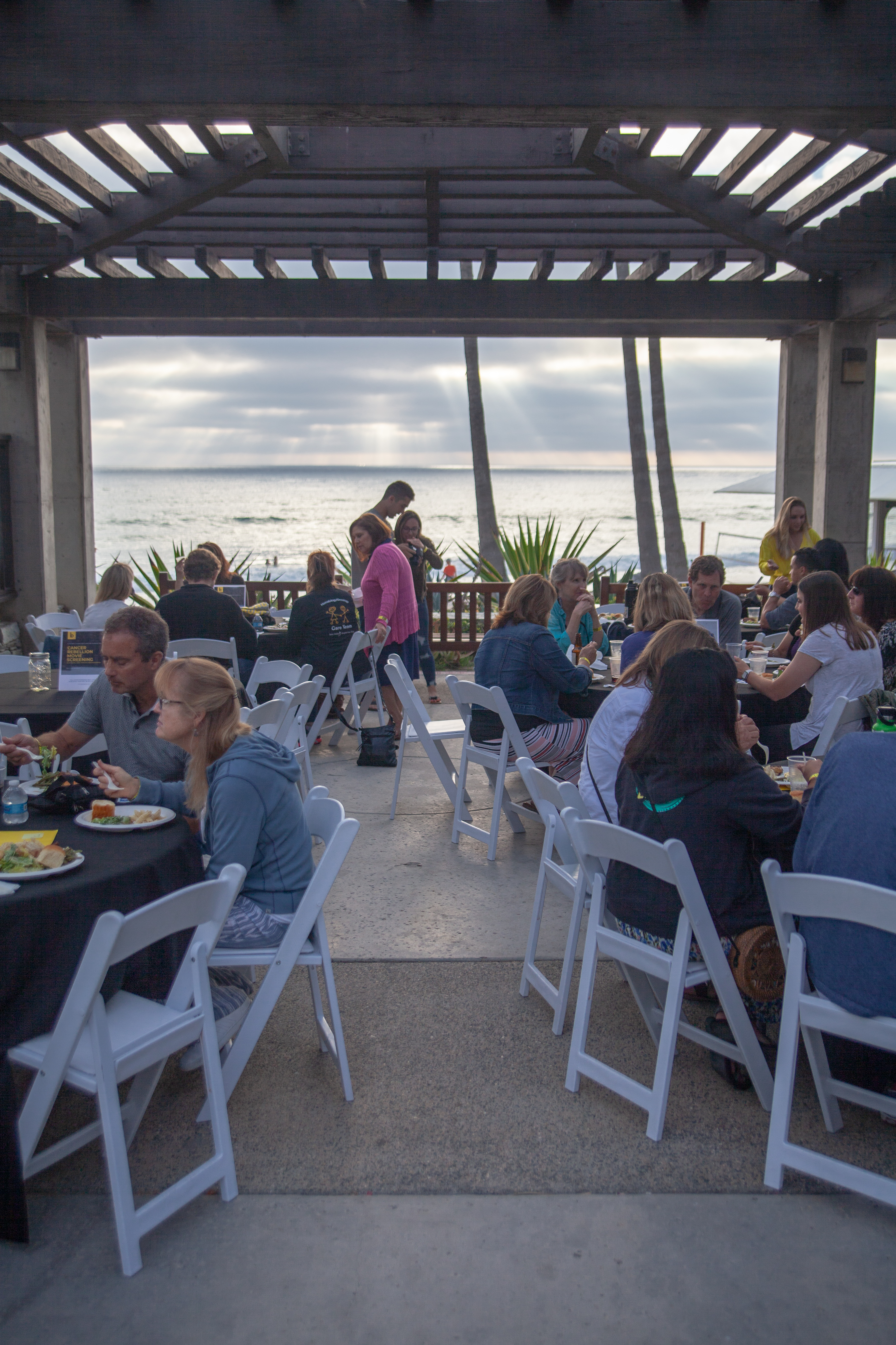People sit at round tables with white chairs, eating and talking under an open-air pavilion by the beach. Sunlight streams through clouds over the ocean in the background. Palm trees and plants are visible near the shore.