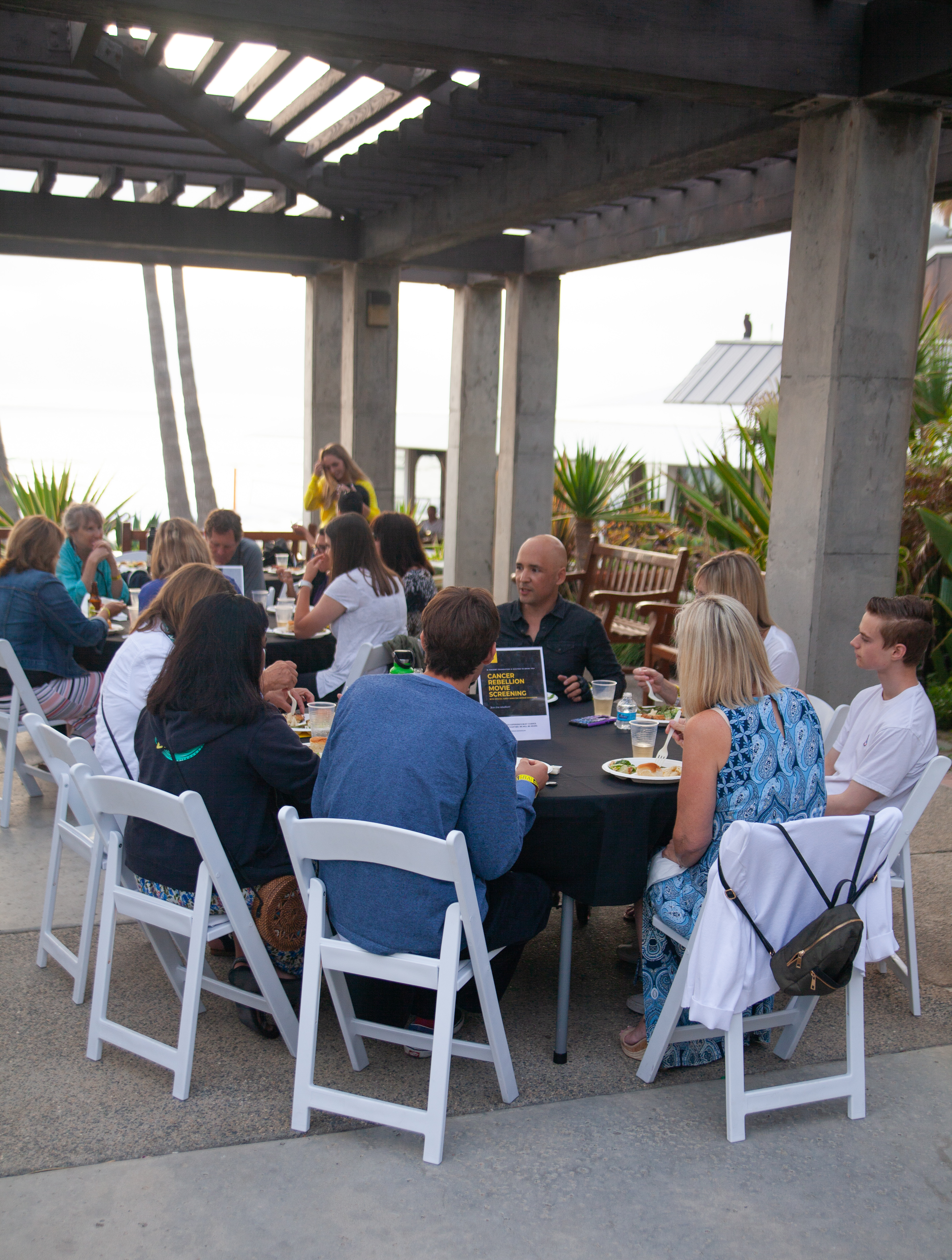 A group of people sit around a round table outdoors under a pergola, eating and talking. Others are nearby, and palm trees and the ocean are visible in the background. It appears to be a social gathering or event.