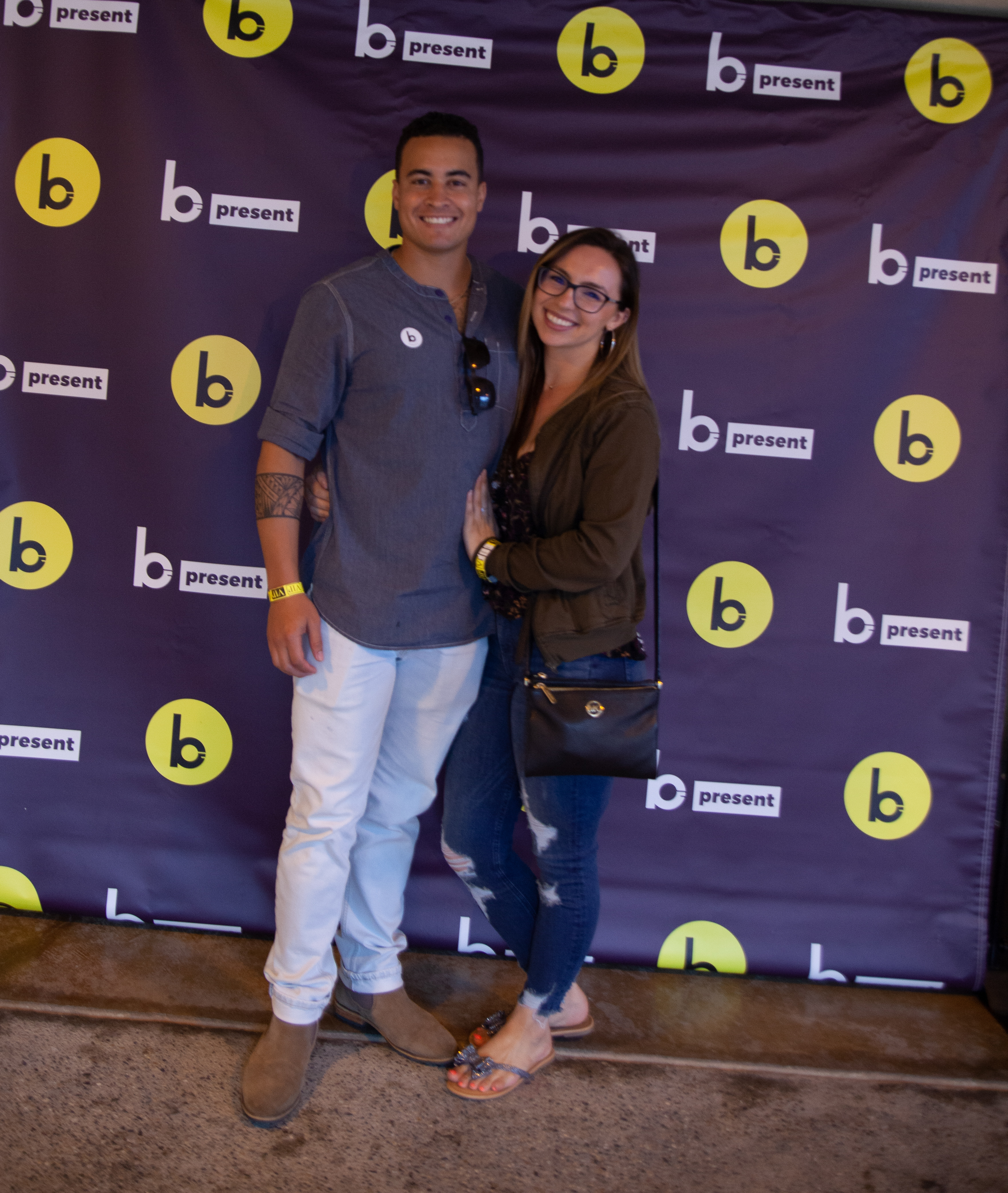 A smiling man and woman stand together in front of a purple backdrop with yellow and white b present logos. The woman hugs the mans waist, and both are casually dressed and wearing wristbands.