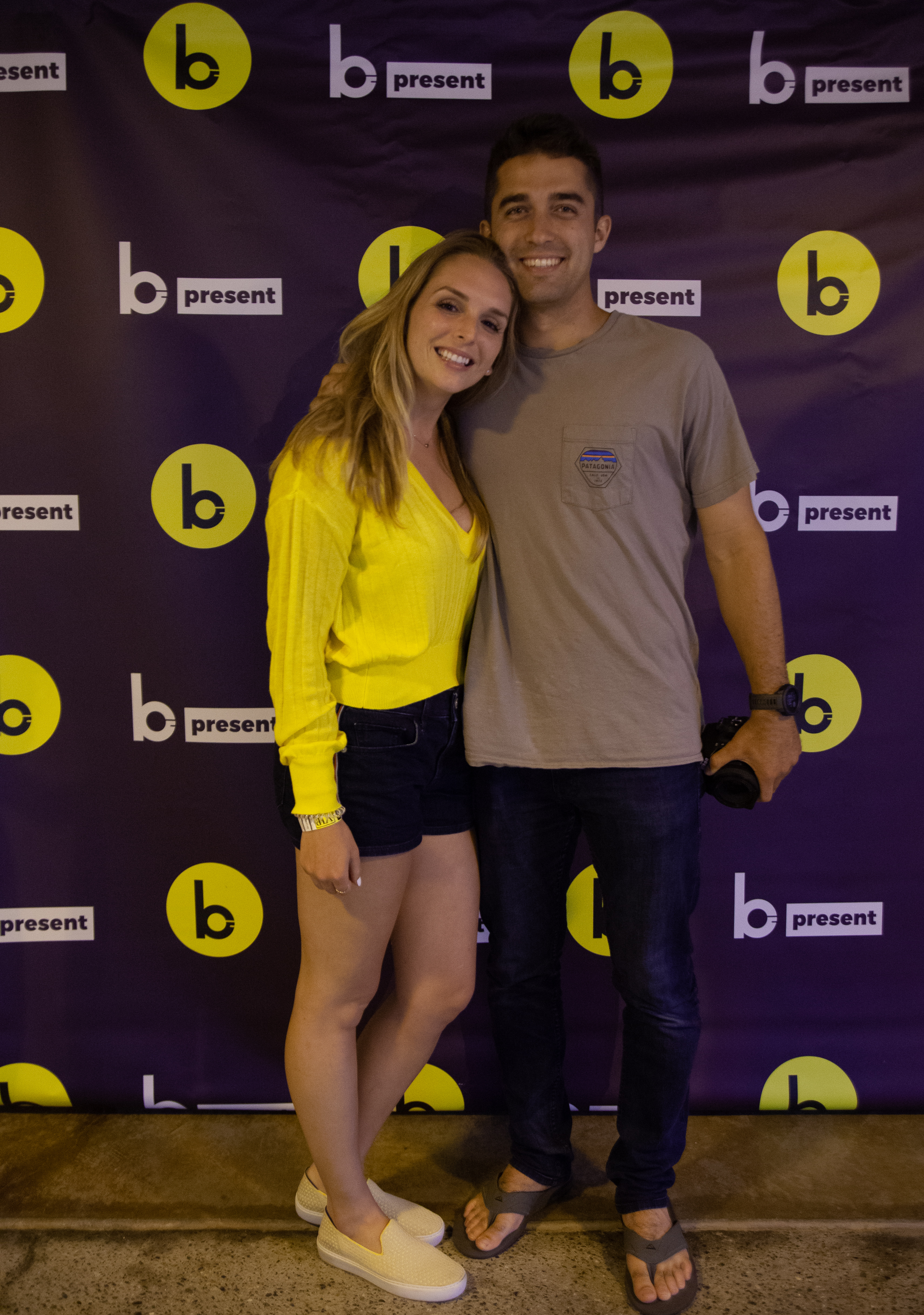 A smiling woman in a yellow top and shorts stands next to a man in a gray t-shirt and jeans. They pose together in front of a purple backdrop with yellow b logos and b present text.