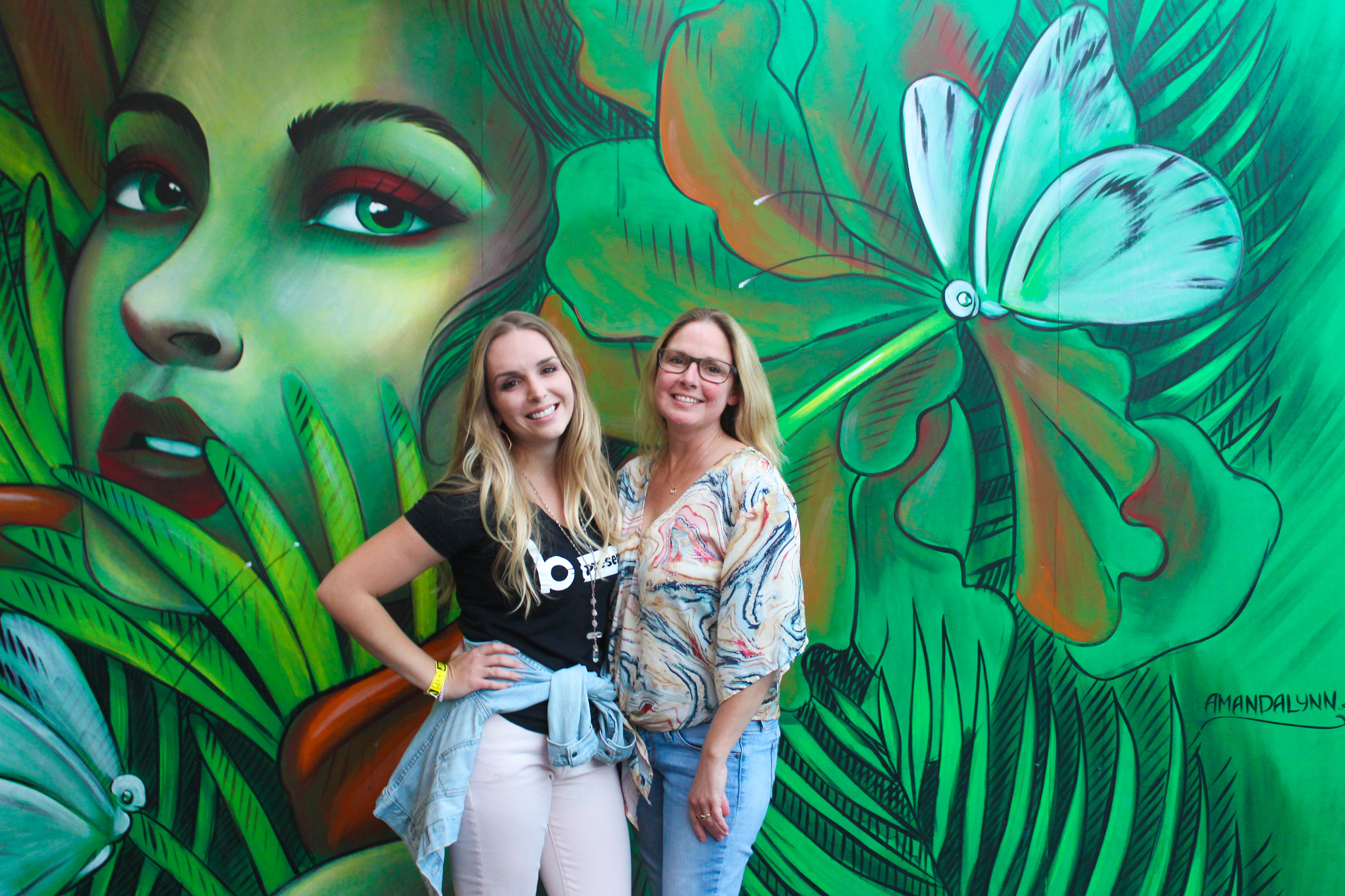 Two women smile and pose in front of a vibrant mural featuring a large female face, green leaves, and a white flower—its colorful backdrop as lively as a night out at dueling pianos. Reds and greens make the scene pop with energy.