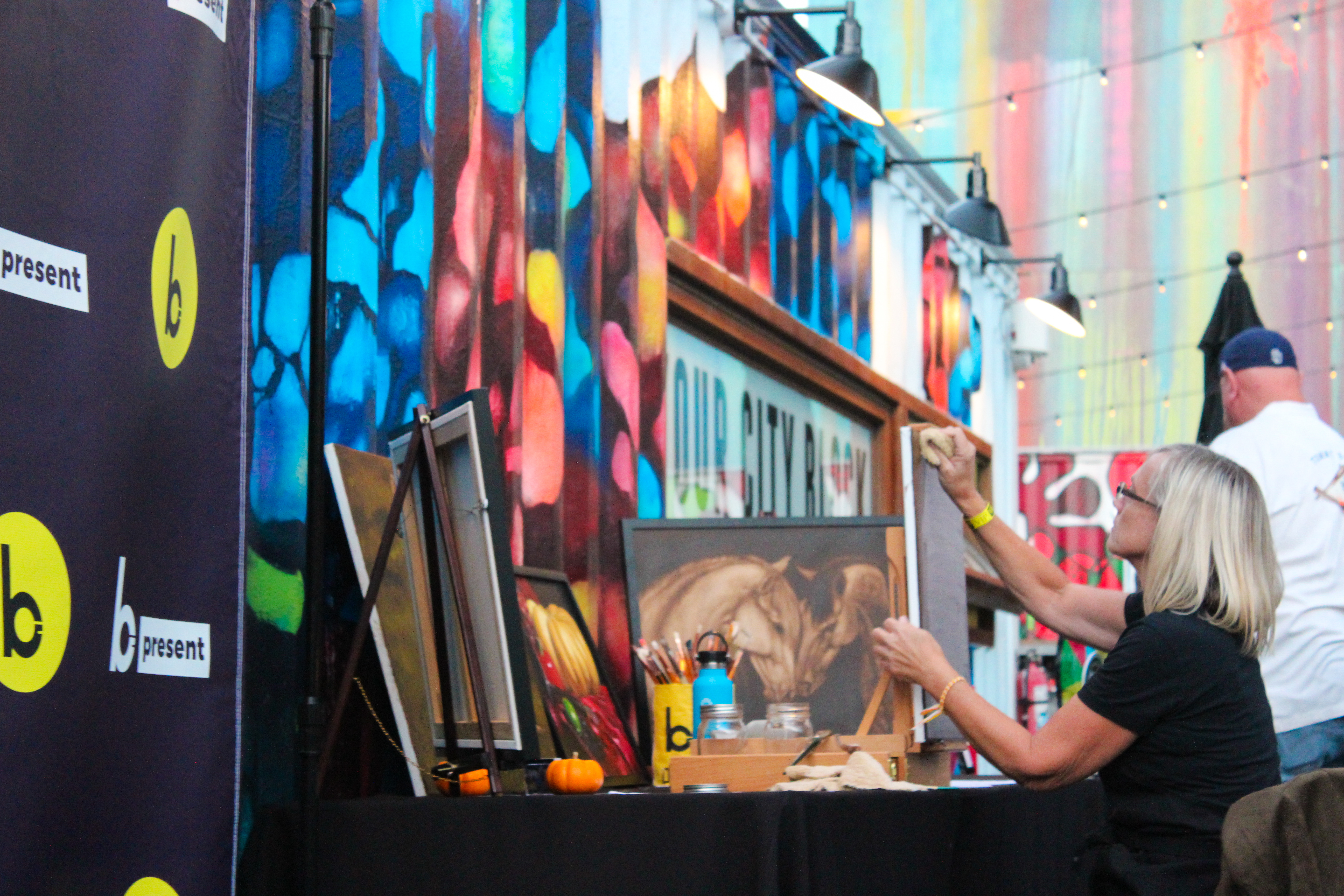 A woman sits at a table painting in front of vibrant, colorful wall art. Several framed artworks and art supplies are on the table. In the background, a man stands nearby as lively as dueling pianos echoing through a creative space.