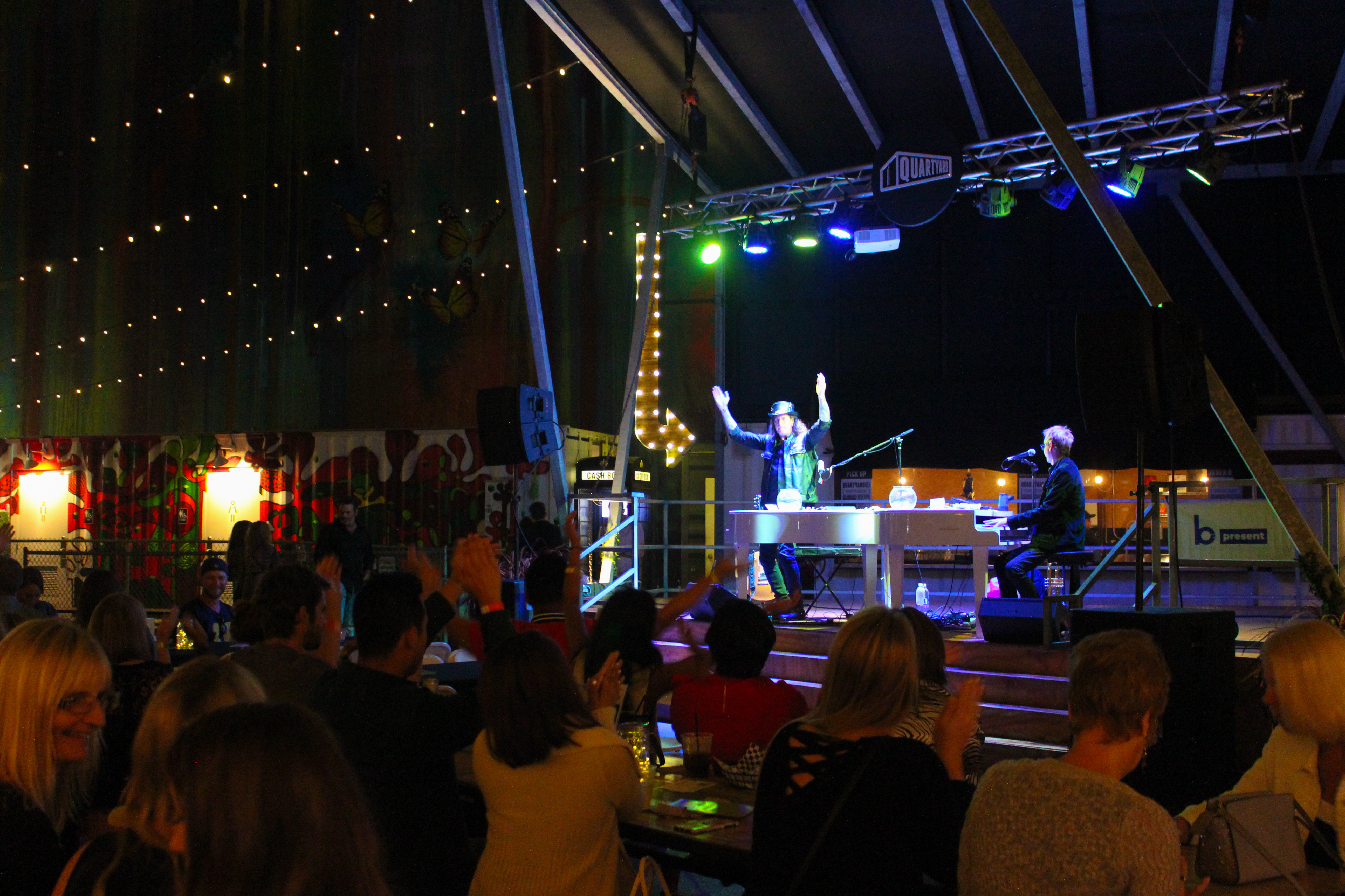 A lively indoor event with a crowd watching a dueling pianos performance on stage under colorful lights. The audience is seated at tables, clapping and cheering, while decorative string lights hang from the ceiling.