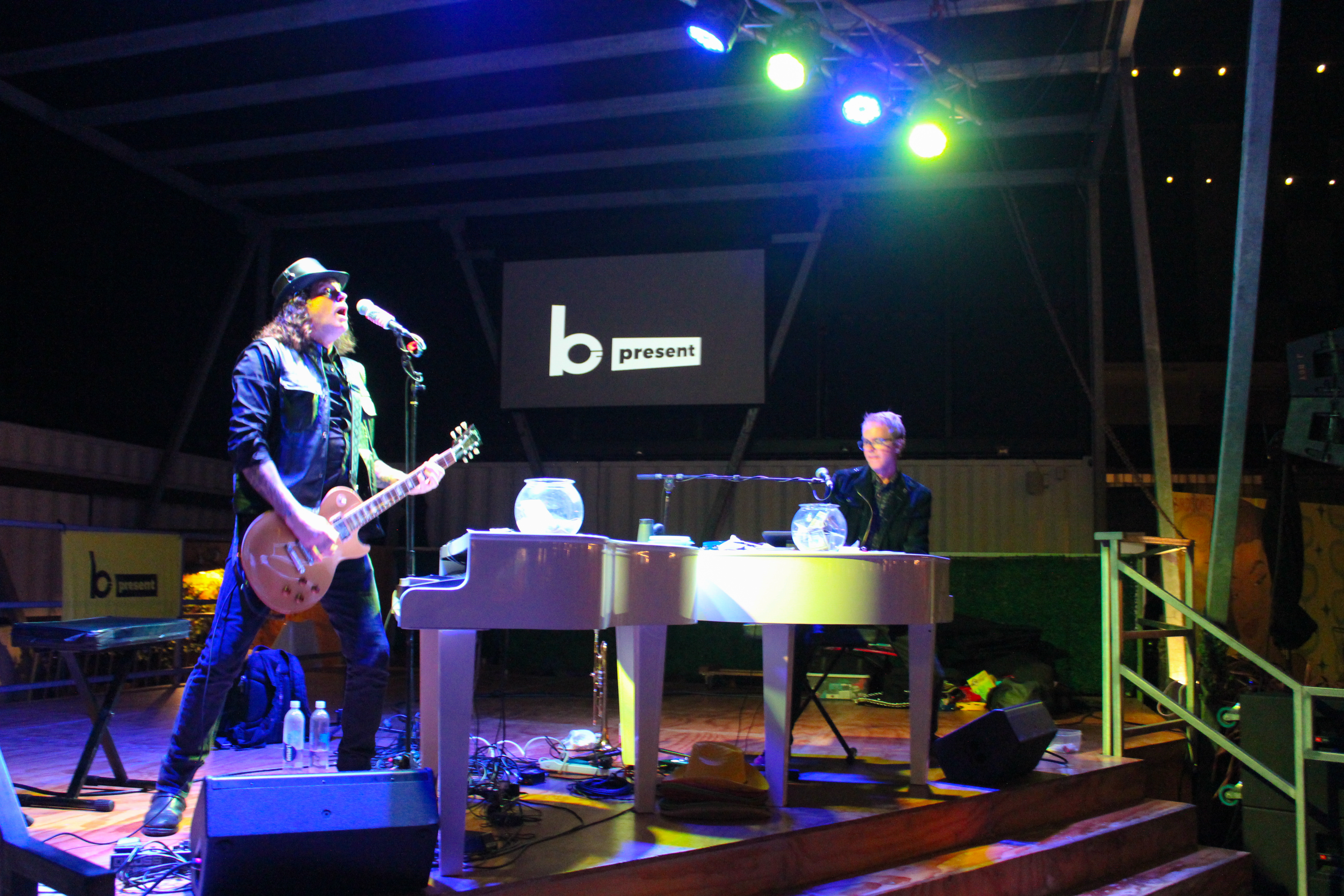 Two musicians perform on stage at night; one plays electric guitar and sings, while the other dazzles on a white grand piano. Bright lights illuminate their dueling pianos, framed by a screen behind them that displays the text “b present.”.