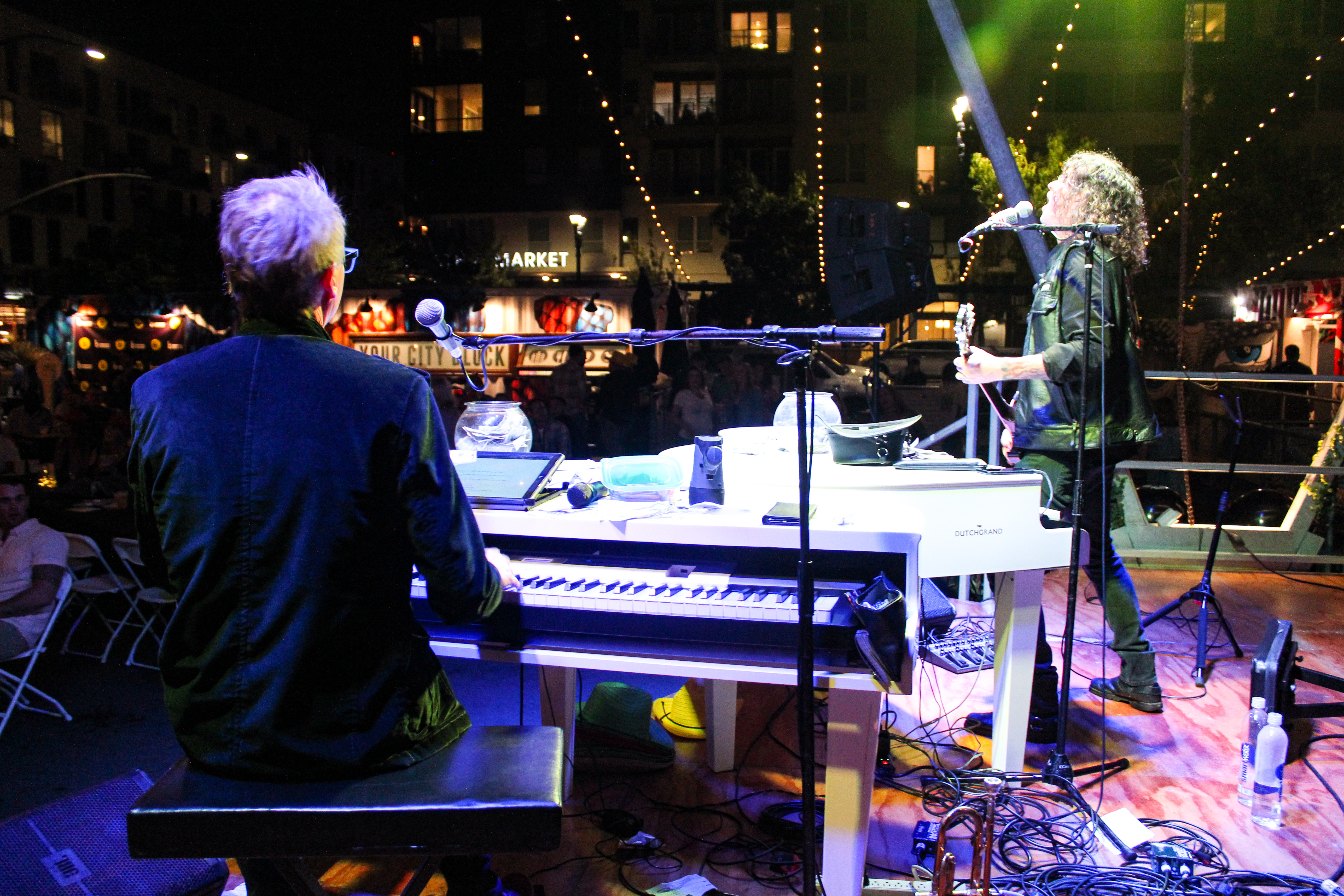 A musician plays a white keyboard on stage while another sings and plays guitar, creating a dueling pianos vibe under colorful stage lights at an outdoor nighttime event, with a crowd and buildings visible in the background.
