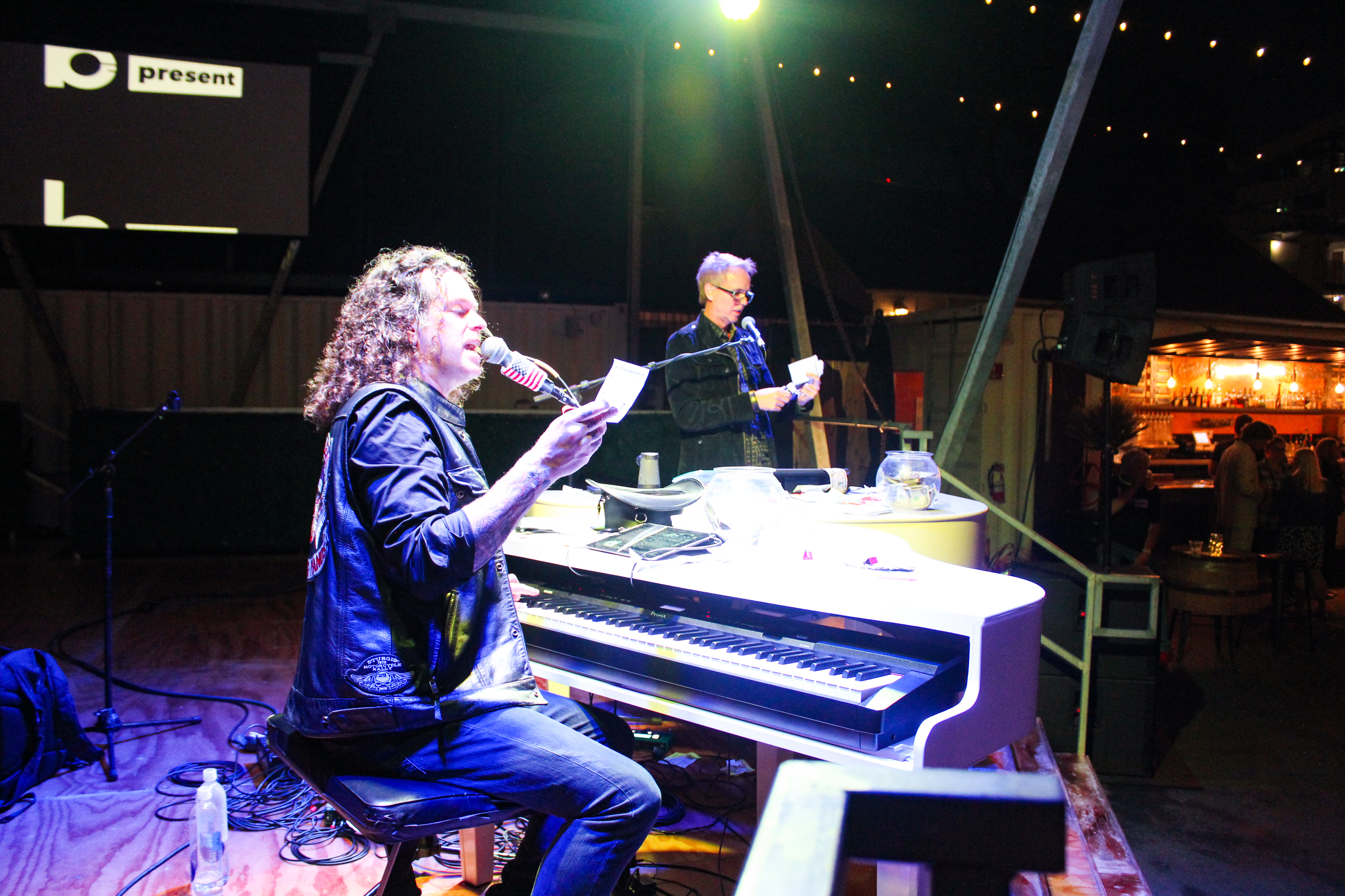 A man with curly hair speaks into a microphone while seated at a white piano on stage during a lively dueling pianos show. Another man stands behind him reading from a paper, as string lights glow above the audience in the dim venue.
