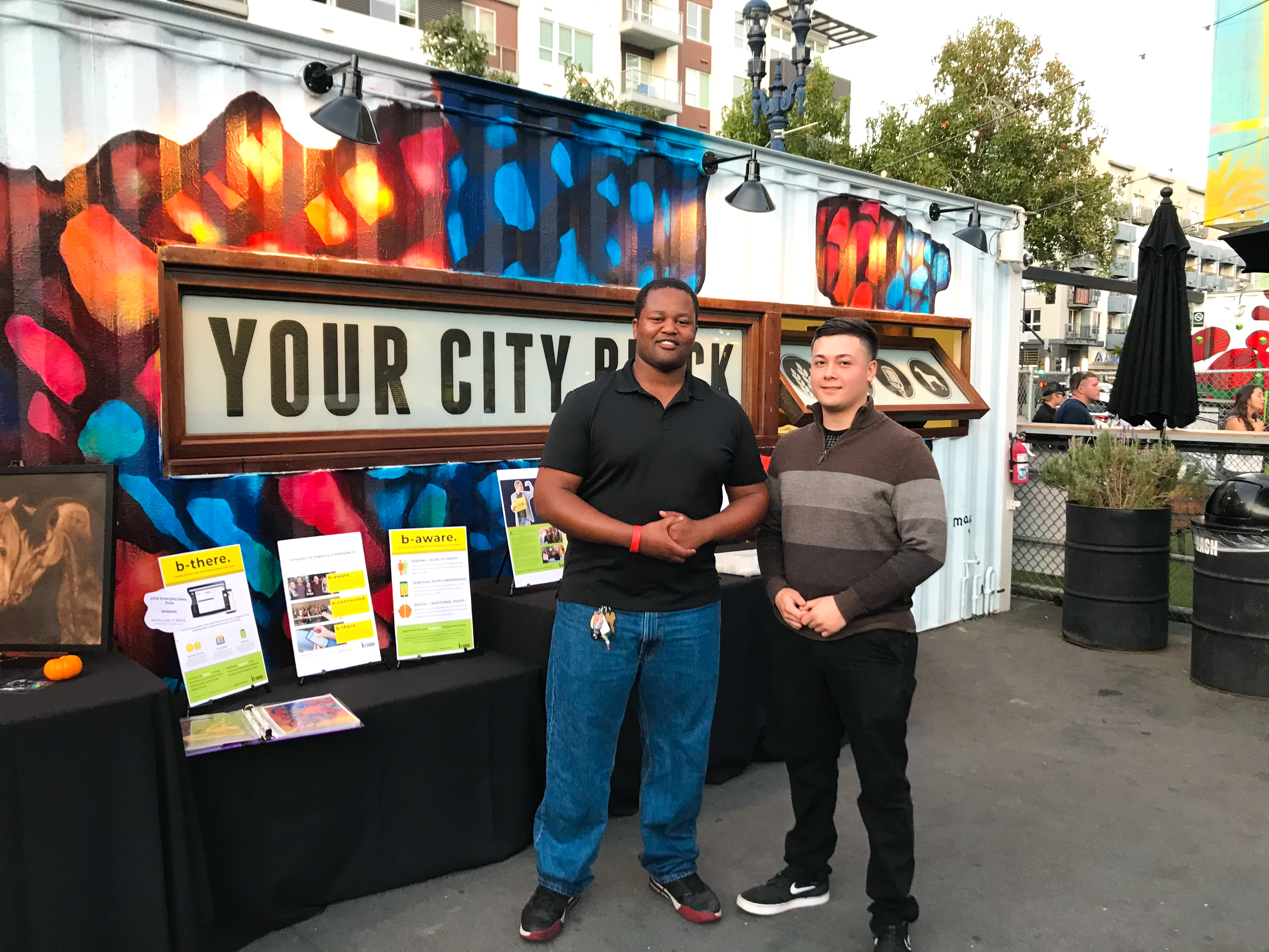 Two men stand smiling in front of a colorful mural that says YOUR CITY BLOCK. At this lively outdoor event—featuring dueling pianos—tables display brochures and signs, with urban buildings and greenery in the background.