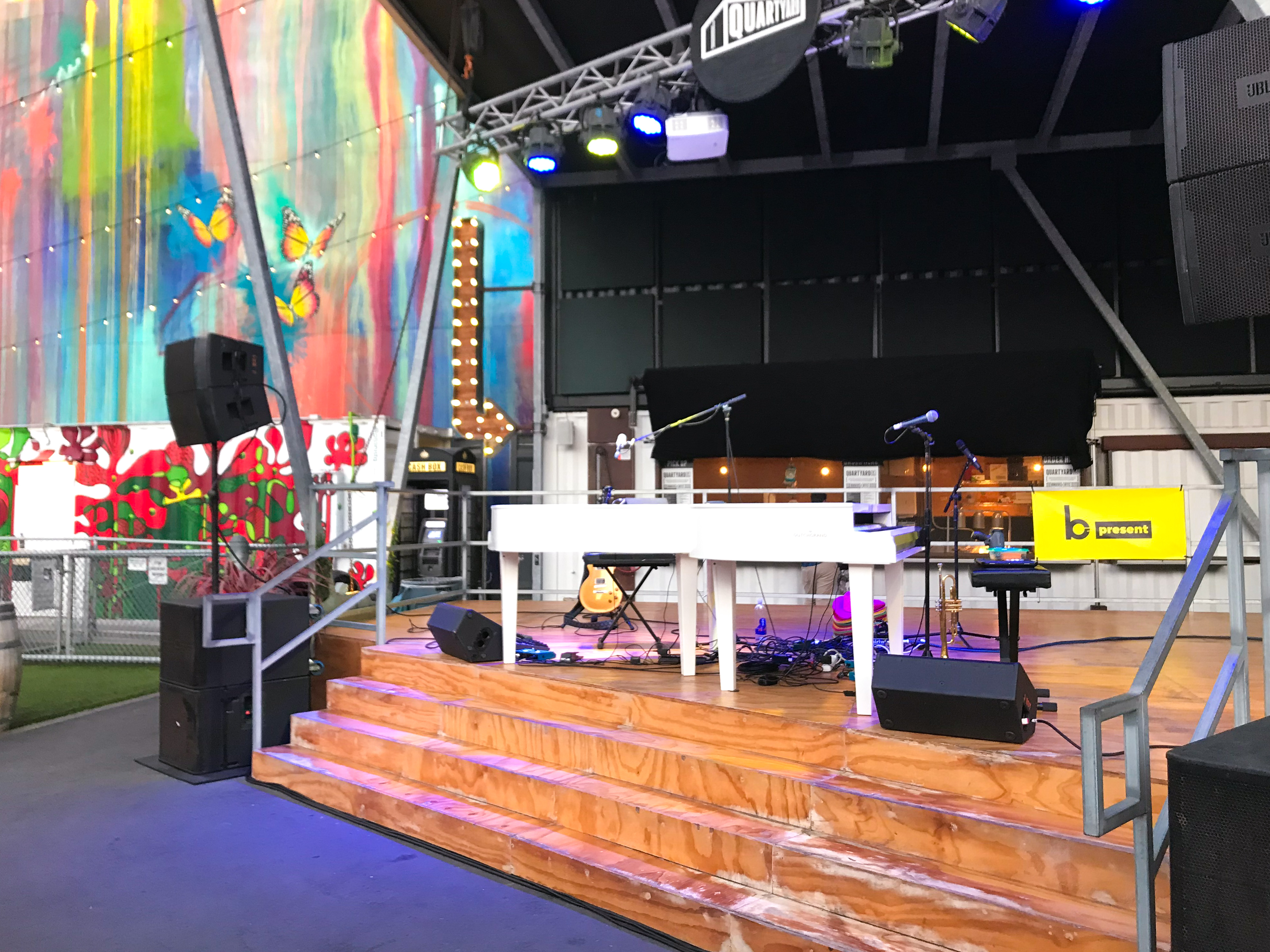 A stage with instruments and microphones set up for dueling pianos, illuminated by colorful lights. Behind it, there’s a vibrant mural featuring butterflies and flowers in an indoor or covered outdoor event venue.