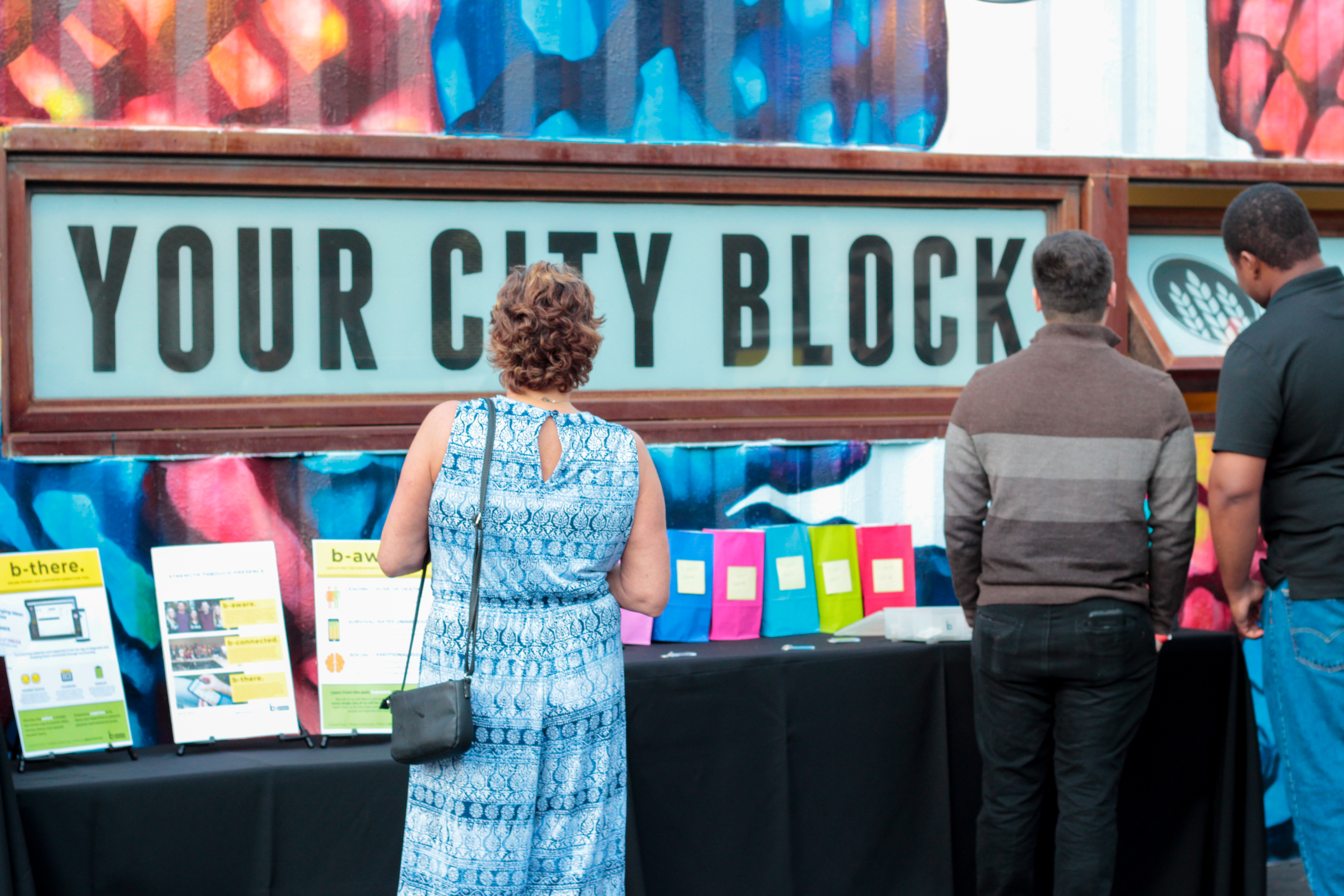 Three people stand in front of a display table with flyers and colorful bags, facing a large sign that reads YOUR CITY BLOCK. The atmosphere is lively—like dueling pianos—with a colorful wall mural adding to the energetic vibe.