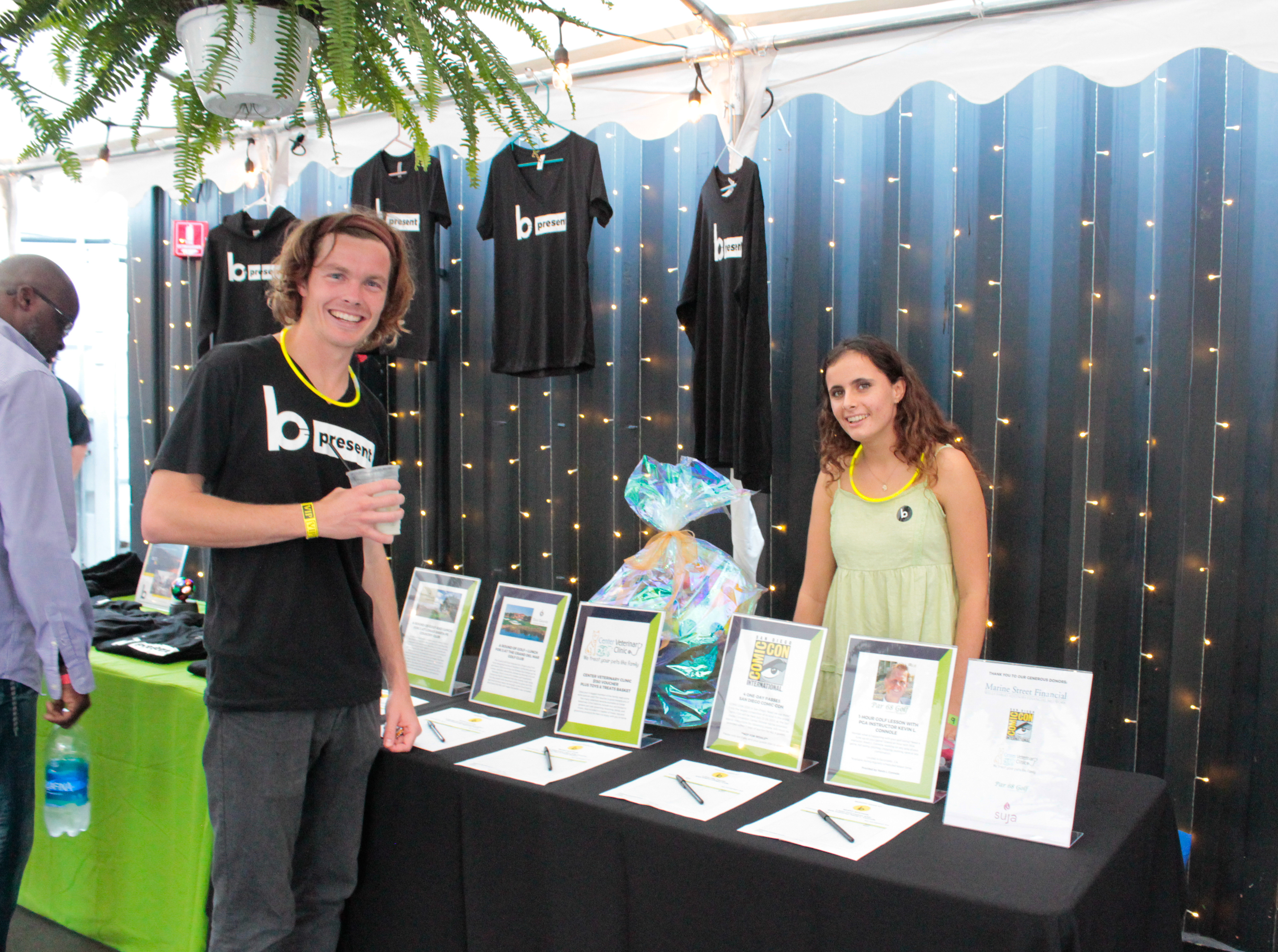 Two people stand and smile behind a black table with framed papers and pens at an indoor dueling pianos event. Behind them, T-shirts hang on a wall with string lights, and a gift basket sits on the table.