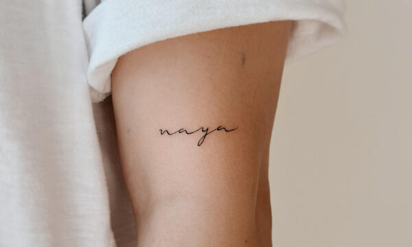 Small fine line script tattoo of the name “Naya” placed on the inner upper arm