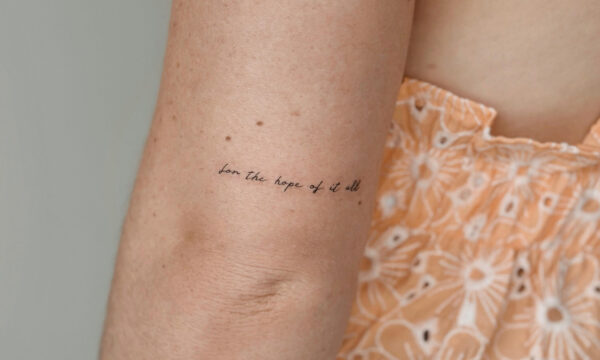 Tiny fine line quote tattoo reading “for the hope of it all” placed on the inner upper arm