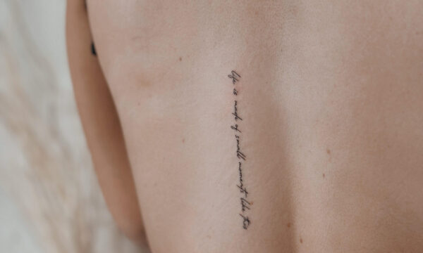 Delicate fine line script tattoo placed vertically along the spine with handwritten quote lettering