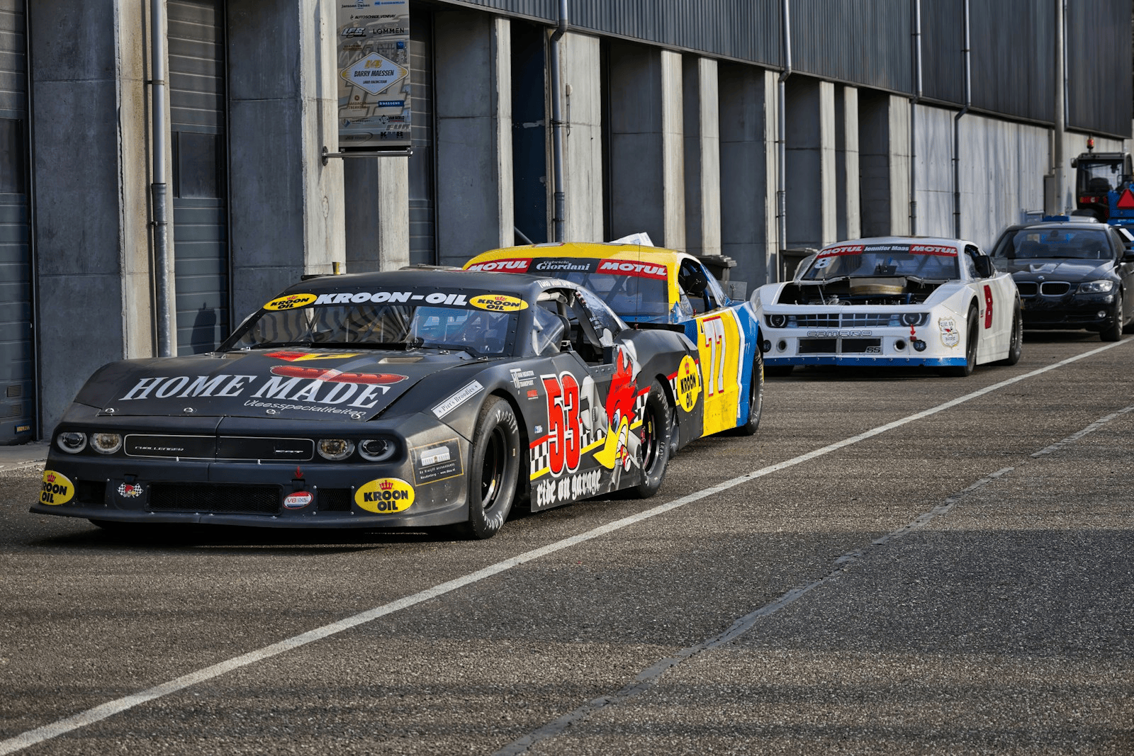Stock cars lined up in pit lane against a concrete garage wall before a short-track race