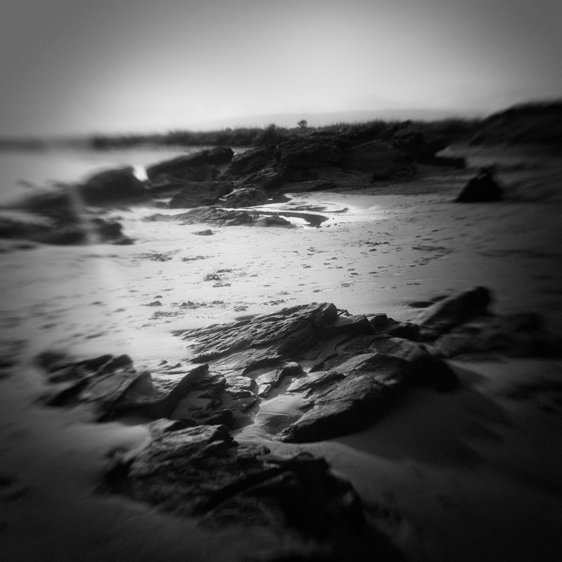 Black and white photo of a flare of light over a sandy beach with dark rocks