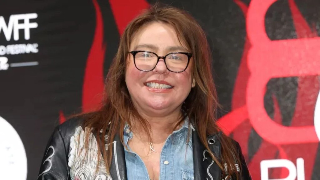 Rachael Ray smiling on a red carpet, wearing a patterned leather jacket over a denim shirt and oversized glasses.