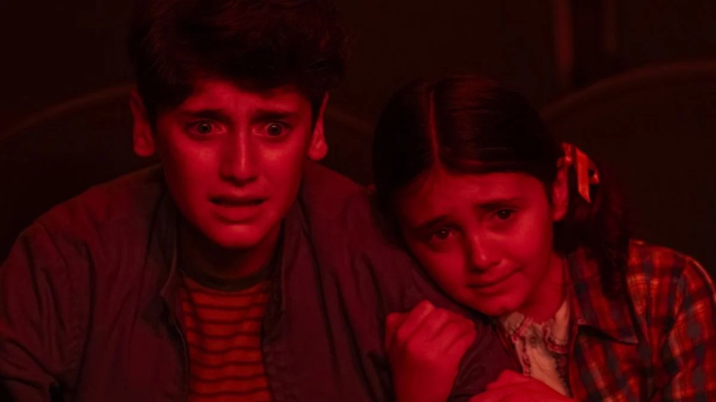 Two children, a boy and a girl, shown in an extreme close-up, illuminated by a deep red light, both screaming or looking terrified in a movie theater.