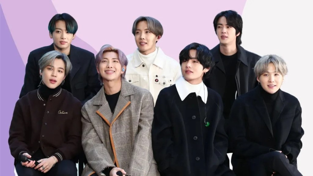 All seven members of BTS sitting together in stylish coats and sweaters against a pink and white background.