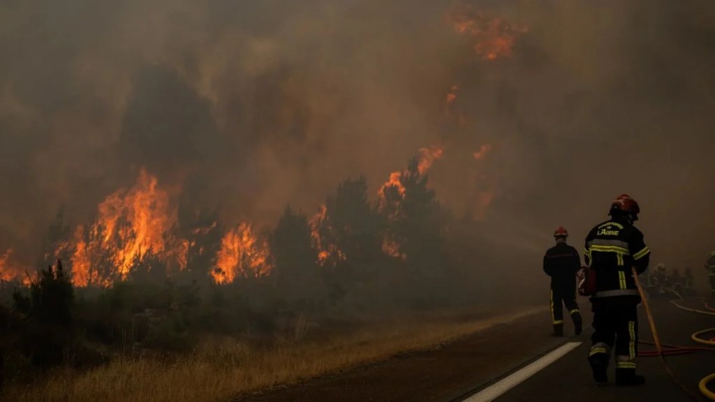 Firefighters in L'Aisne uniforms walk along a smoke-filled road as massive flames devour a pine forest during the 2026 France wildfire emergency.