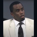 Music mogul Sean Diddy Combs wearing a white suit coat and dark tie while speaking into a microphone.