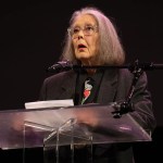 Canadian poet and classicist Anne Carson standing at a podium wearing glasses and a dark blazer while speaking at a microphone.
