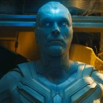 Paul Bettany as White Vision with glowing blue eyes and a mind gem forehead emblem sitting in a dimly lit, dusty room.