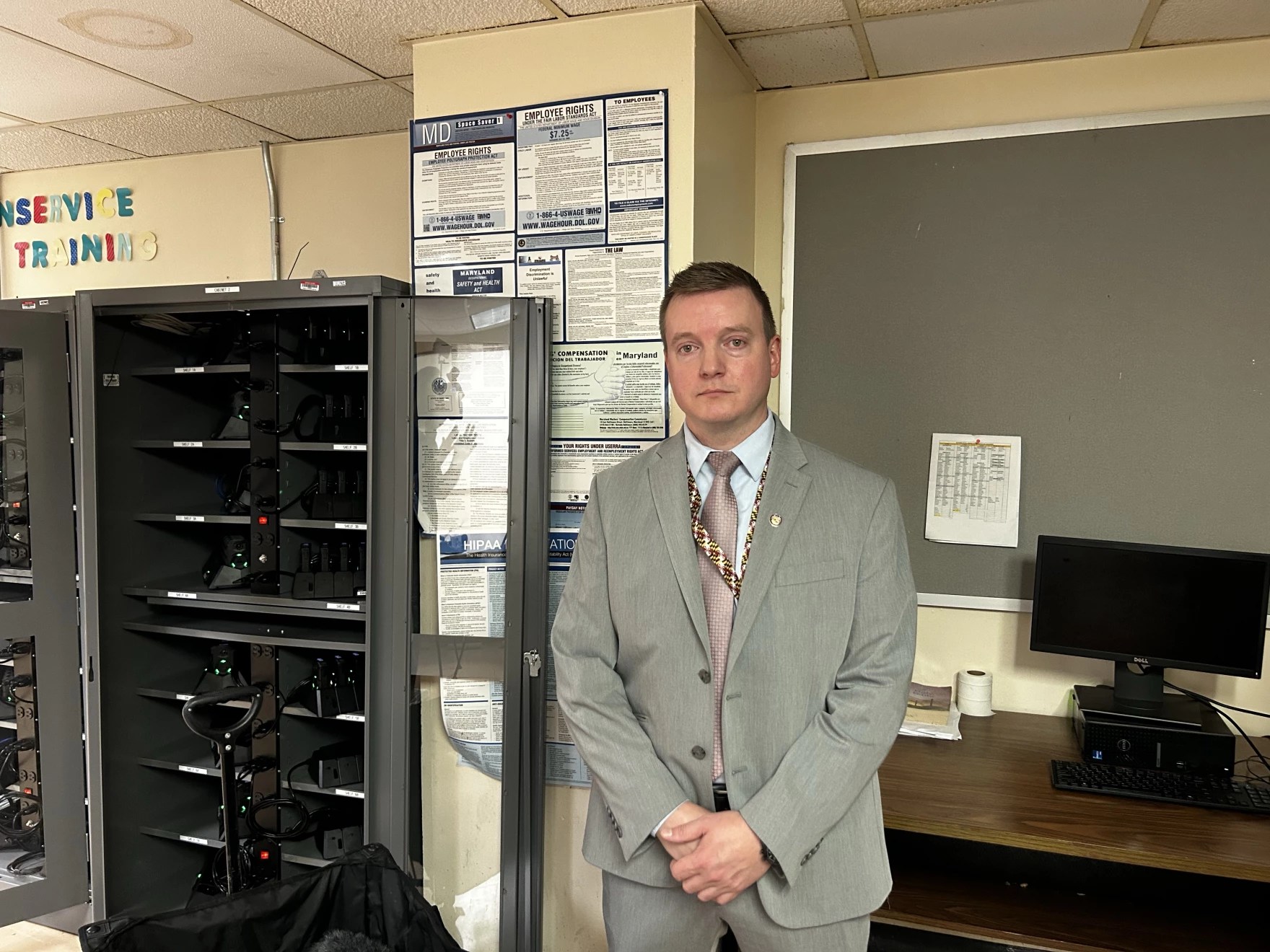 A man in a gray suit stands next to a storage cabinet filled with body cameras.