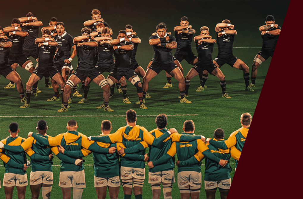 New Zealand rugby players wearing black uniforms perform a Haka, a traditional dance of New Zealand's Indigenous Māori people. South African rugby players in green and yellow uniforms stand next to one another in a line with their arms crossed behind each other's backs.