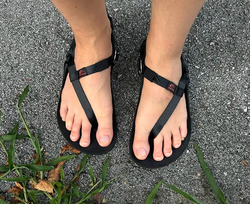 Top view of Bosky barefoot sandals with black straps and toe loop, worn on feet standing on asphalt with grass nearby