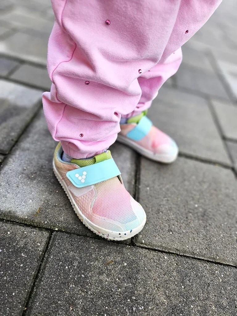 Child wearing brown Vivobarefoot barefoot sneakers with Velcro straps outdoors on grass and wood chips
