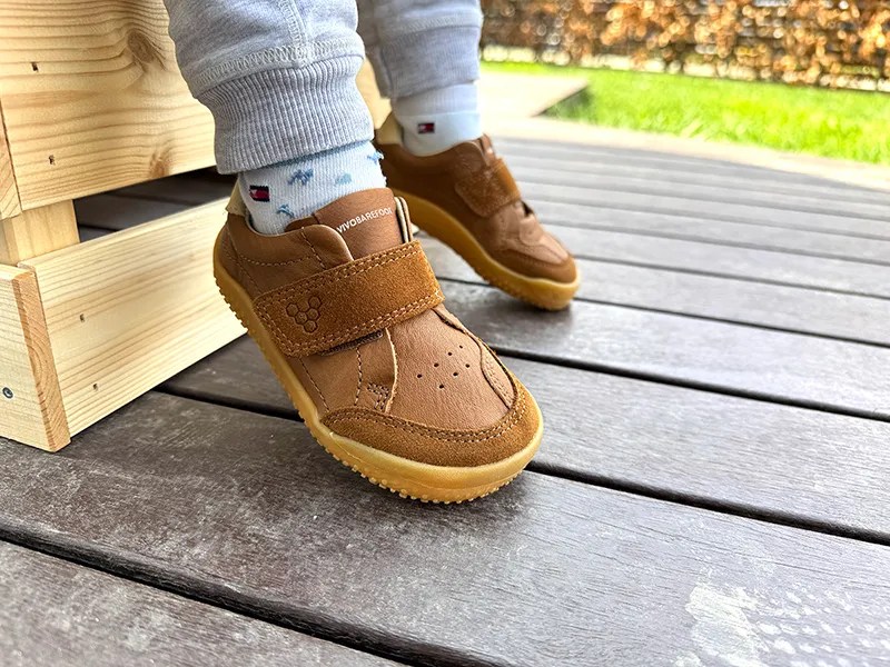 Child wearing brown Vivobarefoot kids sneakers with Velcro straps on wooden deck