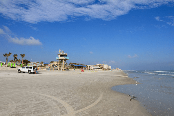 A sunny wide-angle view of a white sand beach in New Smyrna Beach. In the center, a light blue wooden lifeguard tower stands near the water, with a white SUV parked nearby. A few people are scattered along the shoreline under a bright blue sky with light, wispy clouds.