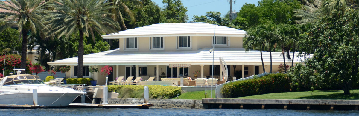 A sunny exterior view of a large, single-story luxury home with a white exterior and a white flat roof. The property is located directly on the water with a wooden dock and a white motorboat moored in the foreground. Lush green palm trees and manicured hedges surround the home under a clear blue sky.