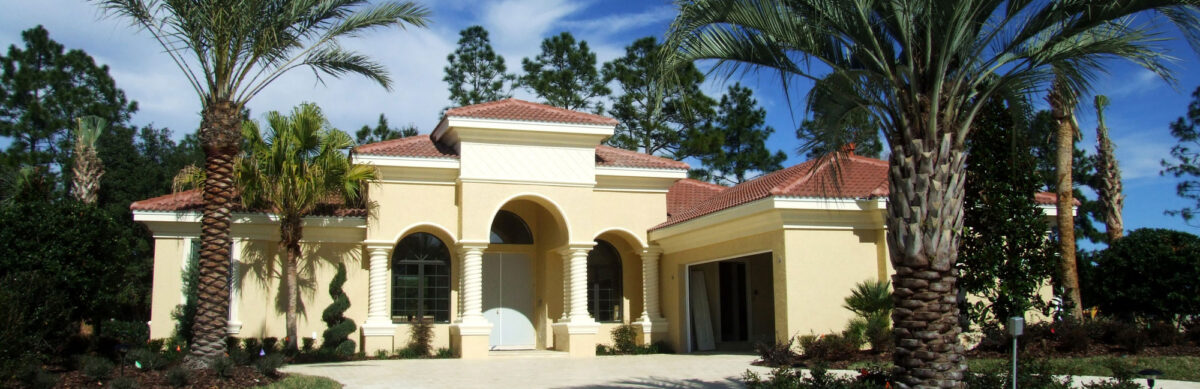 A sunny exterior view of a large, single-story luxury home with cream-colored walls and a terracotta tile roof. The property features a prominent arched entryway, multiple arched windows, and lush landscaping including several tall palm trees and manicured spiral evergreens under a clear blue sky.