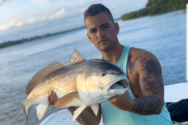 A man with tattoos on his arm, wearing a light blue tank top, poses on a boat while holding a large, golden-scaled red drum fish. A calm river and green treeline are visible in the background under an overcast sky.