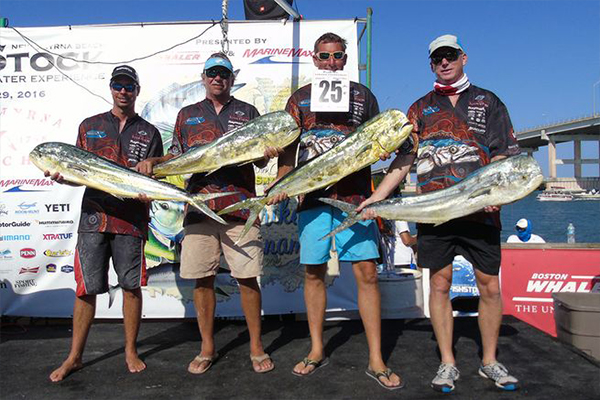 Four men standing in a row outdoors, holding three large mahi-mahi fish. They are posed in front of a tournament banner with MarineMax and Boston Whaler logos, with a bridge visible in the sunny background.