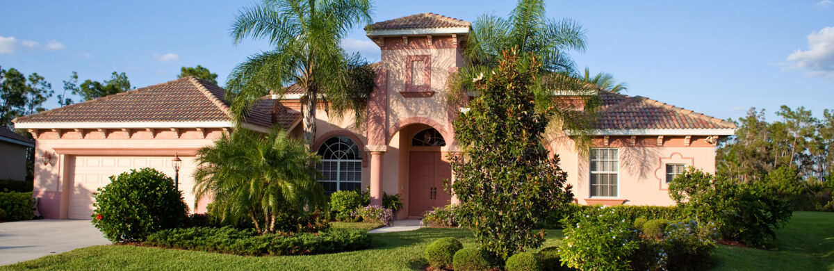 A sunny exterior view of a large, single-story peach-colored luxury home with a terracotta tile roof and white arched accents. The property features an arched entryway, a three-car garage, and lush landscaping with palm trees and manicured bushes under a bright blue sky.