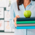 Teacher holding stack of textbooks with green apple on top in classroom setting.