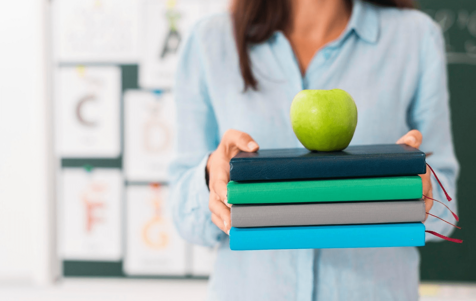 Teacher holding stack of textbooks with green apple on top in classroom setting.