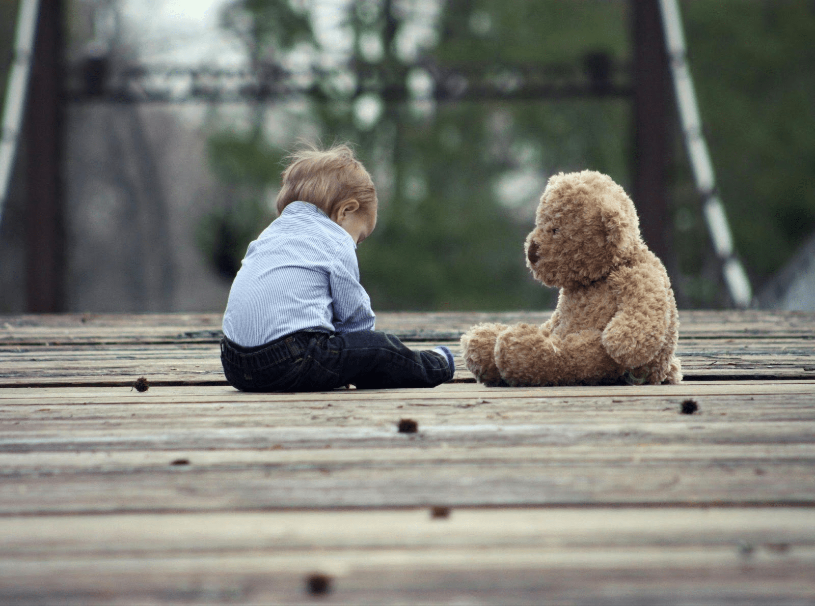 Young child in striped shirt sitting on wooden dock next to brown teddy bear.