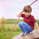 Young boy in red shirt smiling while playing on wooden playground equipment with rope, outdoors in grassy field.