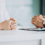 Two people's hands clasped on white table during professional meeting with clipboard and pen visible.