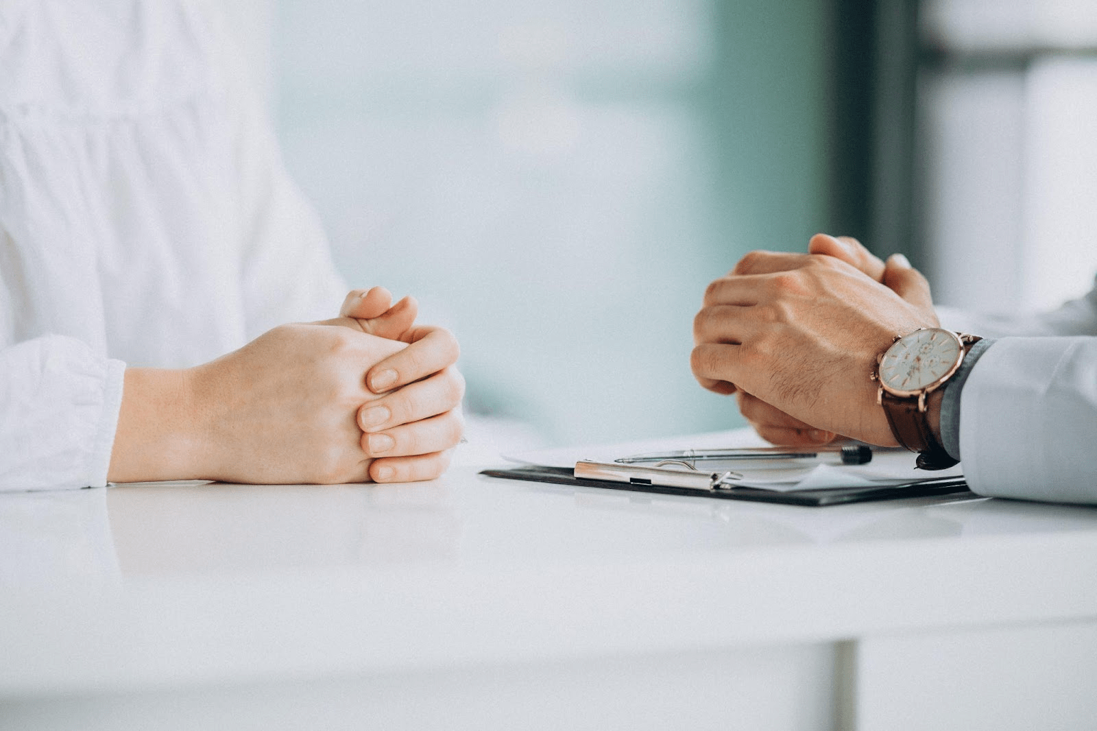 Two people's hands clasped on white table during professional meeting with clipboard and pen visible.